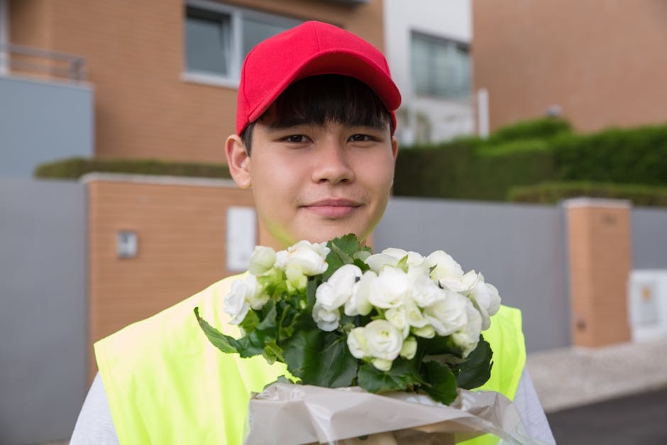 A young male delivery person wearing a red cap and a high-visibility yellow vest is holding a floral gift consisting of a bouquet of small, white roses with green leaves. The flowers are wrapped in wh