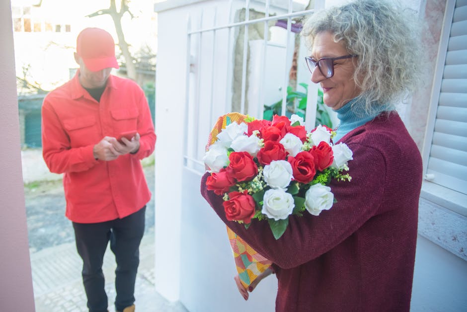A woman with curly gray hair, wearing glasses and a maroon sweater, holds a vibrant bouquet of fresh red and white roses wrapped in colorful paper, positioned near a glass door interior. Outside, a pe