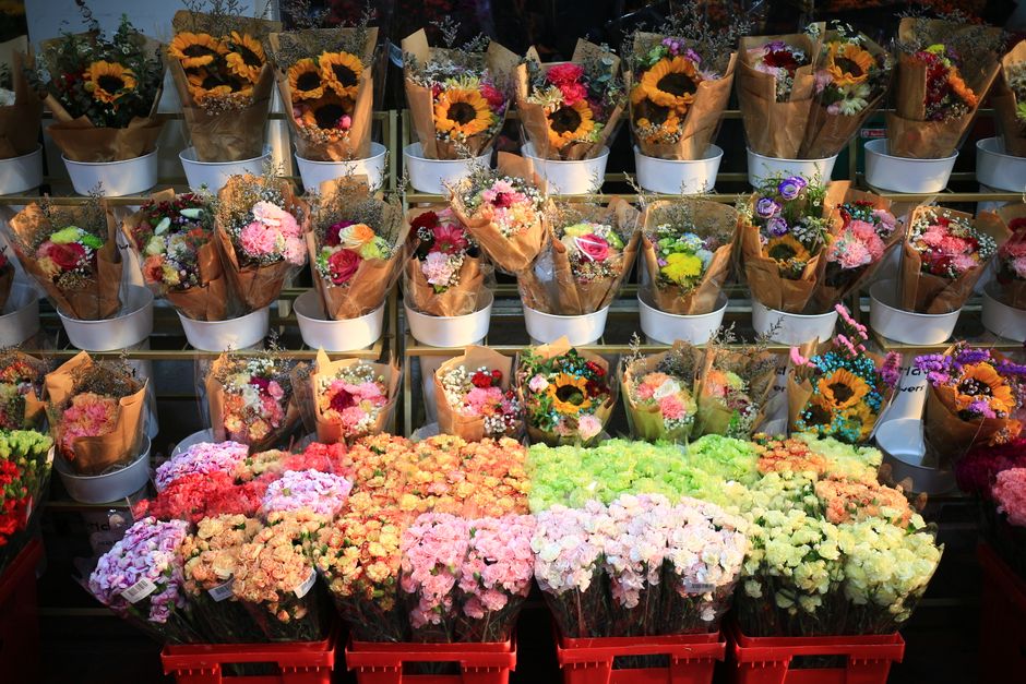 A large selection of fresh flower bouquets arranged in white and brown paper wrappings, featuring vibrant sunflowers, pink roses, white carnations, and multicolored small blooms. The bouquets are neat