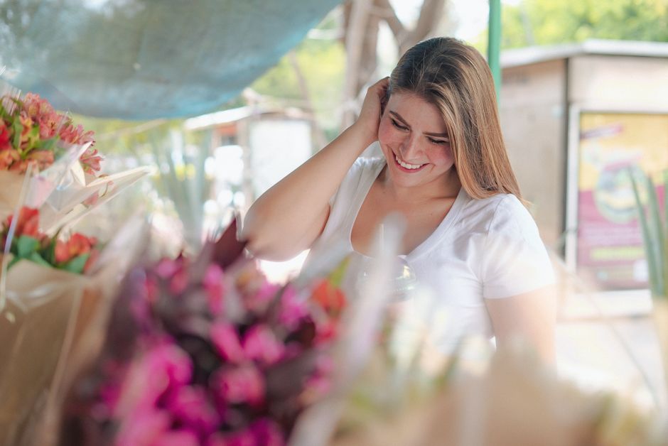 A smiling woman with long brown hair and fair skin, wearing a white t-shirt, is surrounded by an assortment of freshly arranged flowers. The floral display includes pink, red, and purple blooms, with 