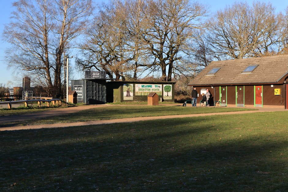 A wide view of Brereton Heath park showing a brown building with colorful vertical panels and a large green sign that reads 'Welcome to Brereton Heath,' set against a backdrop of tall, leafless trees 