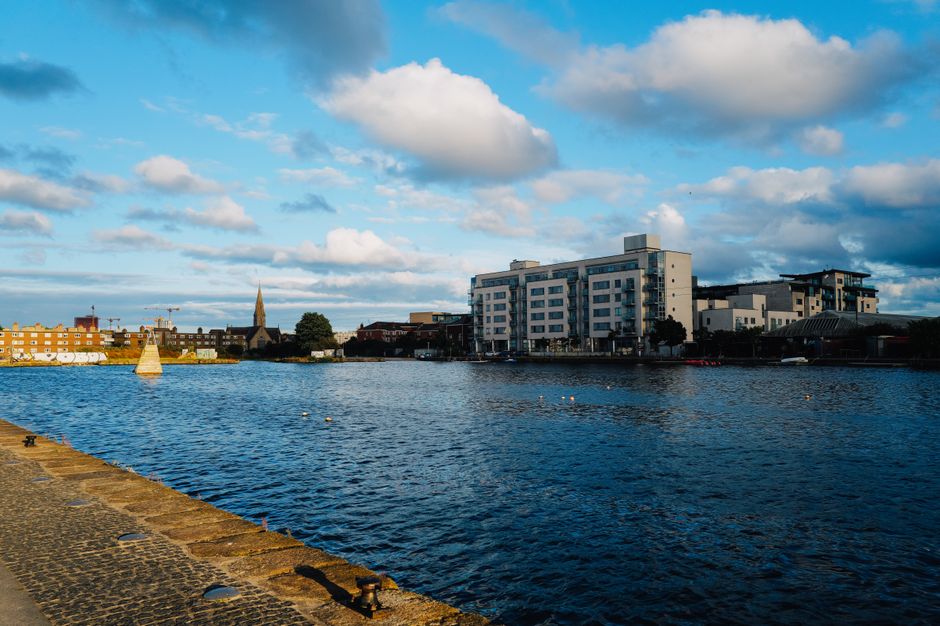 A panoramic view of the Erith Riverside featuring calm river waters reflecting the partly cloudy sky with large white and gray clouds. In the foreground, a stone embankment lines the riverbank, leadin