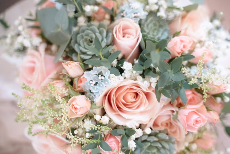 A close-up view of a floral bouquet featuring soft pink roses, small pale pink spray roses, and delicate white baby's breath, accented with silvery-green eucalyptus leaves and succulent plants. The fl