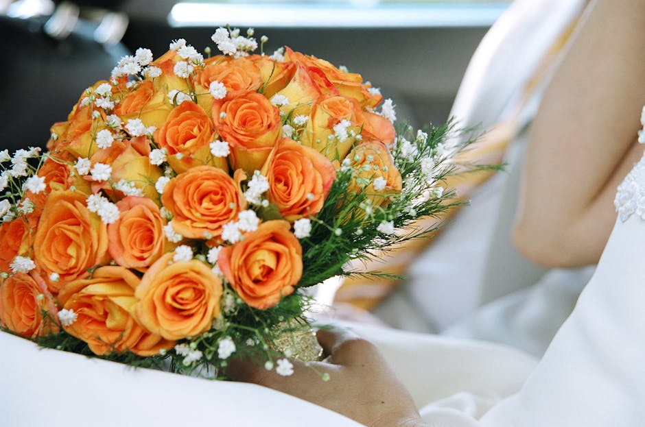 A vibrant floral bouquet featuring tightly arranged orange roses with velvety petals, accented by small white filler flowers and sprigs of greenery. The bouquet is held by a person dressed in white, p