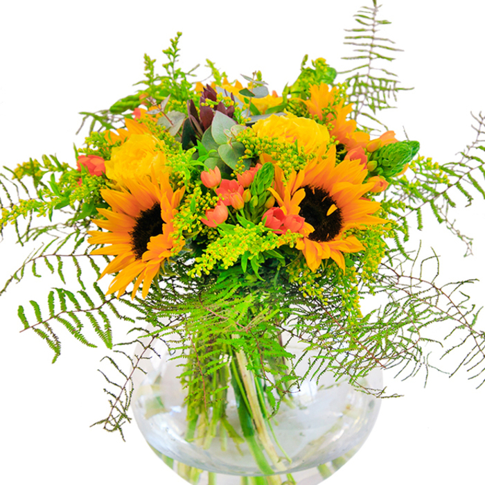 The first impression of this floral masterpiece is unmistakably one of warmth and exuberant colour-an expertly arranged bouquet, crafted in Erith, combining sun-gold sunflowers and soft yellow roses that appear to radiate light from within. Each sunflower's broad petals reach outward in a jubilant display, while the tightly layered swirl of the garden roses offers a refined and elegant counterpoint. Proudly positioned in a clear, rounded glass vase evocative of classic designs found in Erith's bustling High Street shops, the bouquet is accented by cascades of deep green leaves and trailing strands of feather-light ferns. Scarlet Hypericum berries shine against the matte backdrop of petals, their plump, glossy forms adding a sparkling dimension to the composition. Among the foliage, gentle sprigs of bluish-green eucalyptus and wisps of yellow-blossomed filler catch the eye, their textures and hues suggesting a fresh harvest from nearby Crossness Nature Reserve. The fresh-cut stems, visible through the gleaming glass, are immersed in water to preserve their vitality and fragrance. If you were to lean in, you might catch the delicate blend of floral scents and the whisper of greenery, redolent of summer meadows. Designed by Erith's skilled florists, this bouquet is perfect for celebrating birthdays, anniversaries, or any occasion that calls for joy and heartfelt brightness-a true centrepiece for Erith homes and a mark of local floral artistry.